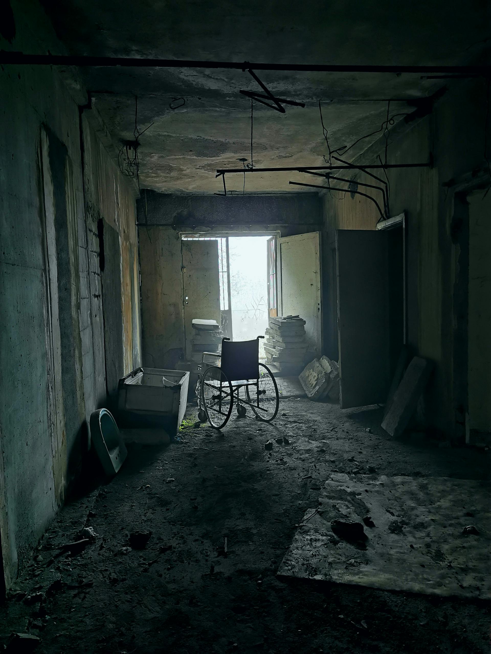 An abadoned hospital hallway featuring broken fixtures, holes in the walls, a broken window, and a broken down wheelchair.