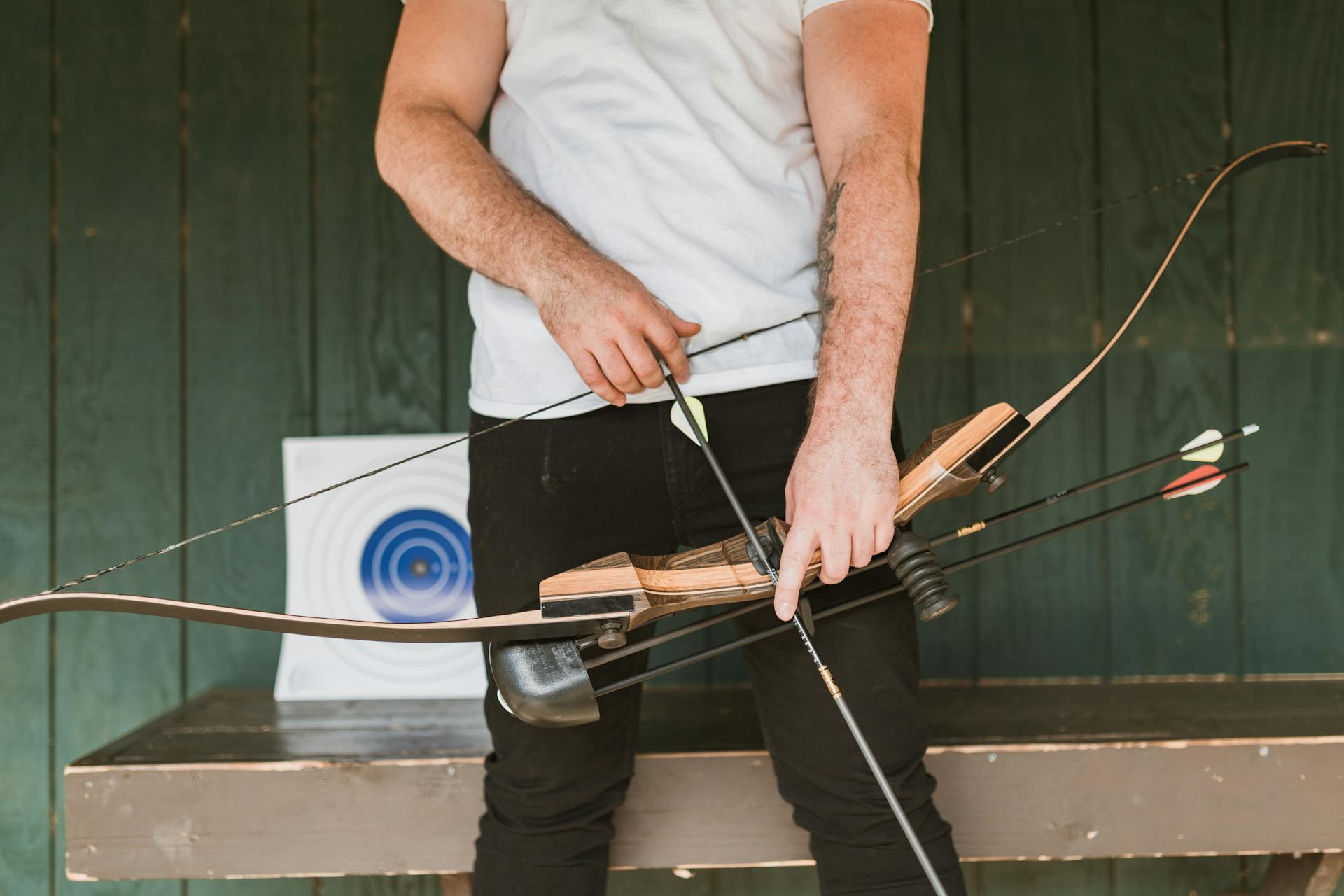 a man wearing a white shirt and black pants holding a hunting bow. He has an arrow in his hands at waist height and it setting up to be shot.