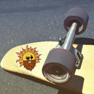 A skateboard with the bottom facing up and a sticker of Remy's sun logo on it. the sticker is of a pop art illustration of a bearded sun with sunglasses winking and smiling.