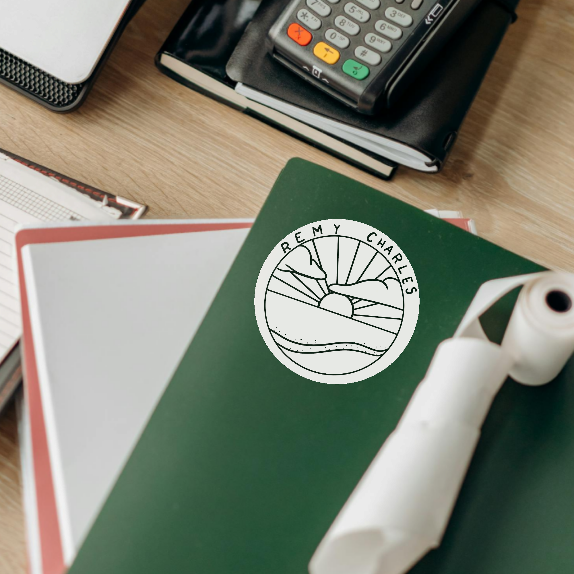 A stack of folders and documents on a desk next to a card processor and accounting items. A green folder displays a sticker of a sunrise or sunset with clouds at a beach in colorless pop art illustration.