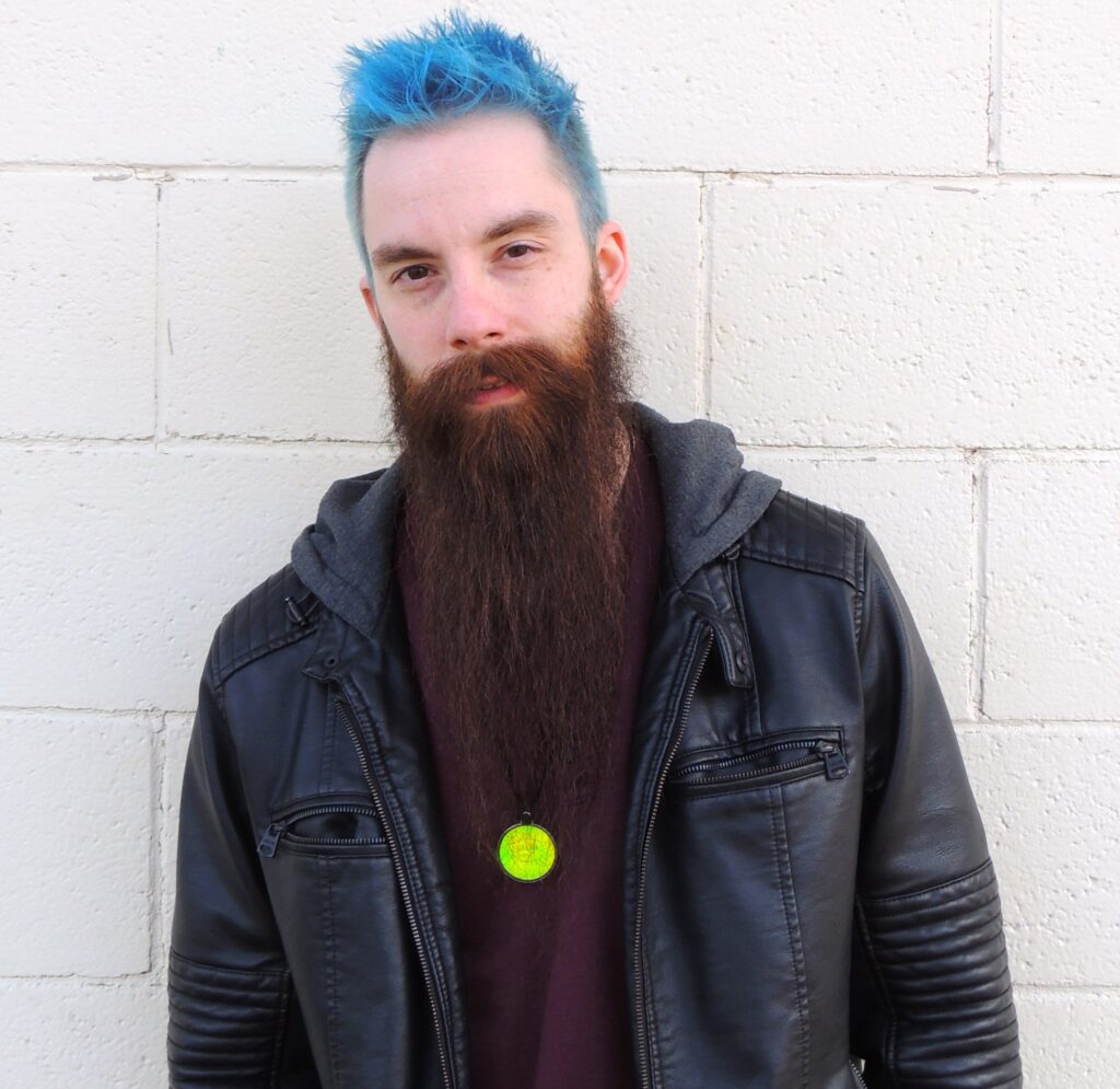 A young Caucasian man with spiked blue hair is staring intensely into the camera. He is standing against a white brick background and is wearing a burgundy shirt, black leather jacket, and magic eye necklace.