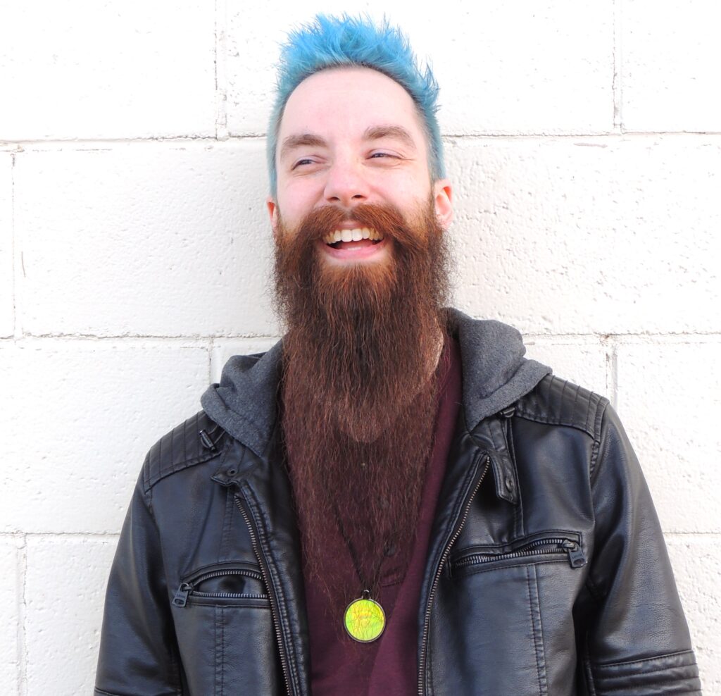 A smiling caucasian man with short spiked blue hair and a long brown beard is standing in front of a white brick wall wearing a burgundy shirt, black leather jacket, and a magic eye necklace.
