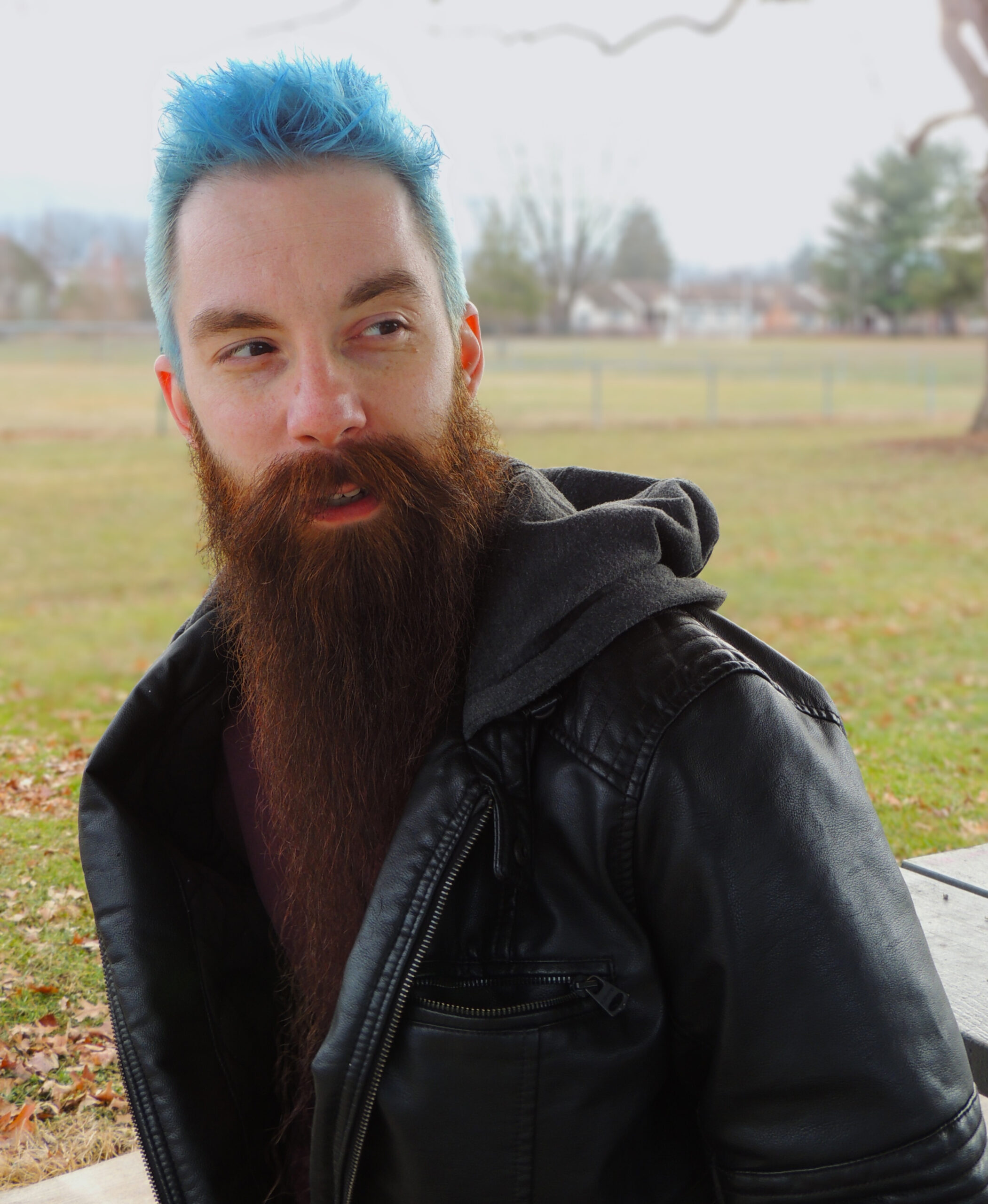 A young Caucasian man with spiked blue hair in a seated position staring over his left shoulder. Behind him are blurred out trees and grass. He is wearing a burgundy shirt and black leather jacket.